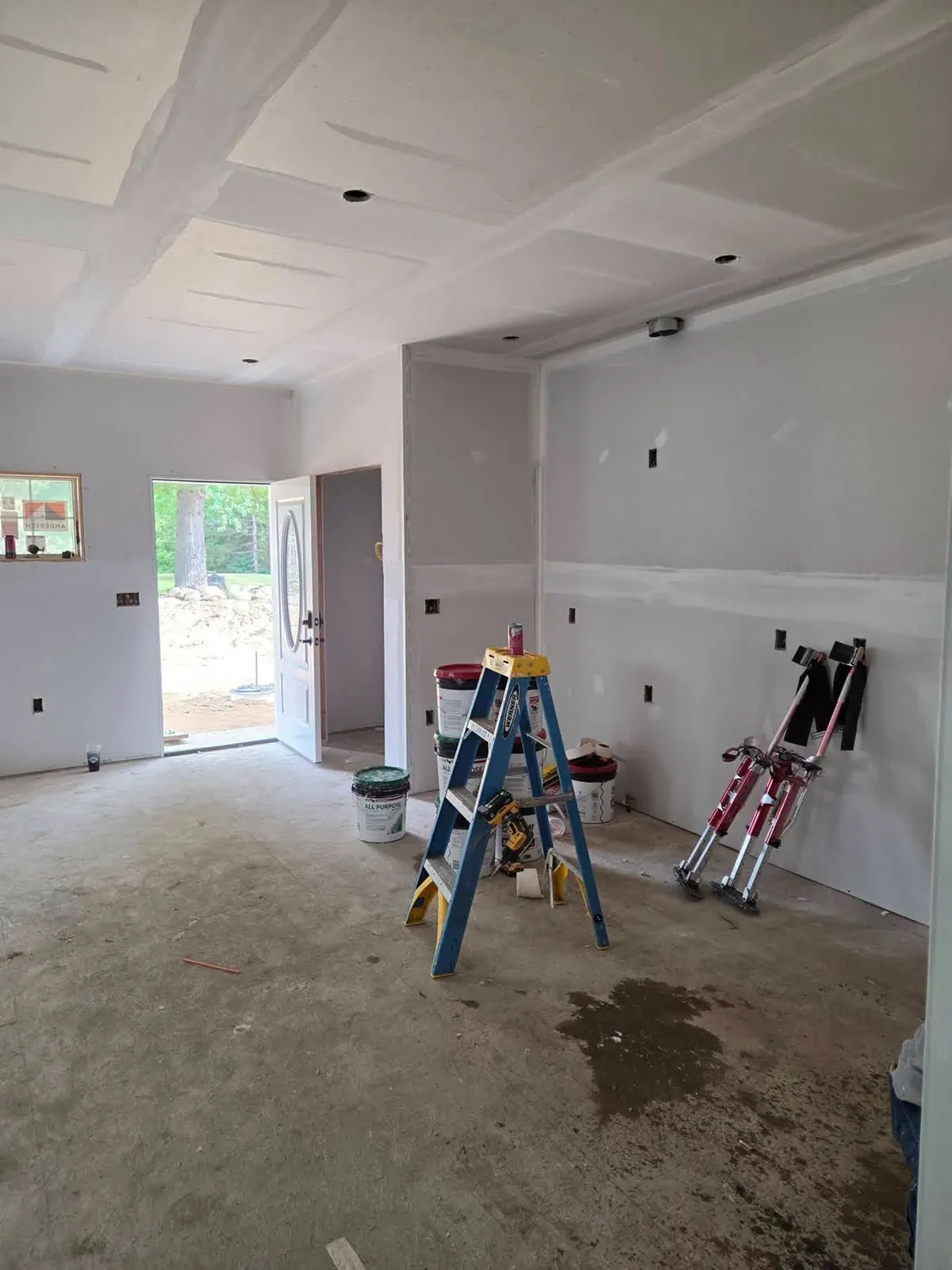 Drywall hung and taped on walls and ceiling, with joint compound buckets, ladder and drywall stilts on site