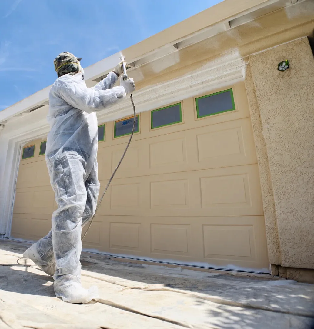 Painter in protective suit spraying exterior paint on a garage door and stucco soffit, with surfaces masked and ground covered