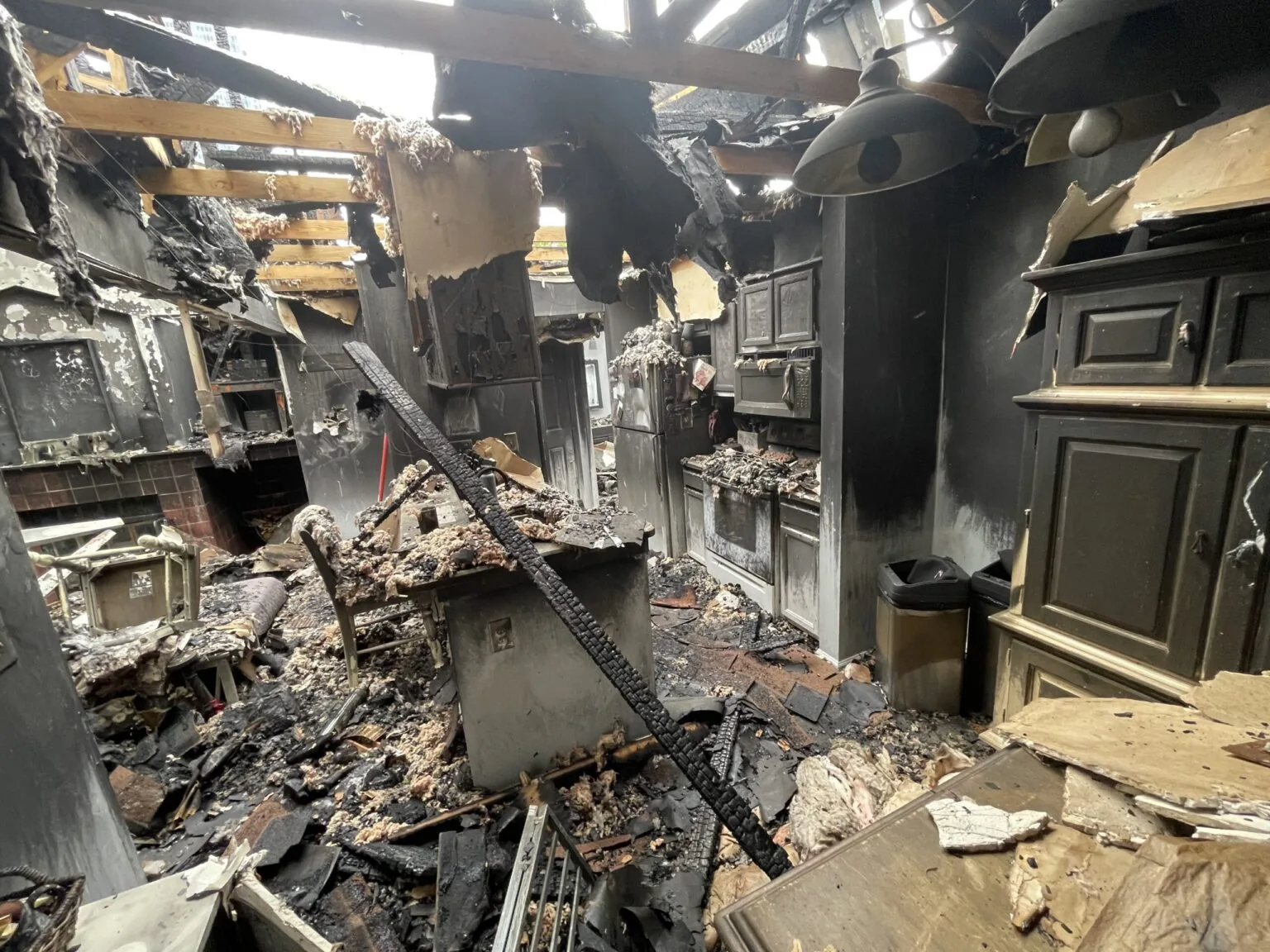 Kitchen and living area after a house fire, with a collapsed roof, charred framing and soot-covered cabinets