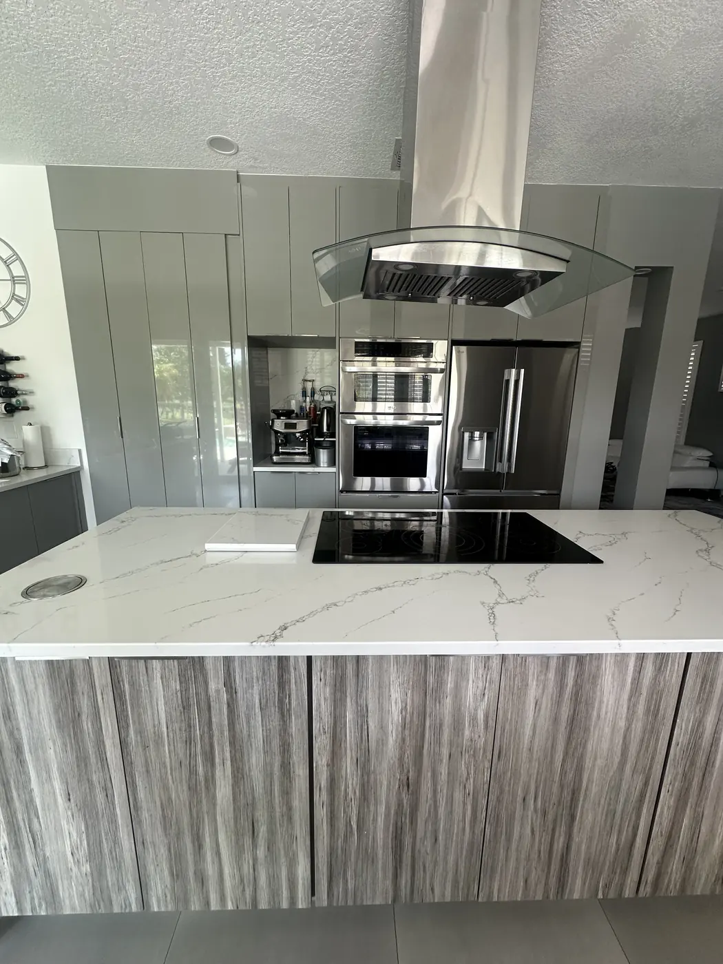 Finished modern kitchen with gray high-gloss cabinets, a wood-grain island with a white veined quartz countertop, cooktop and glass island hood
