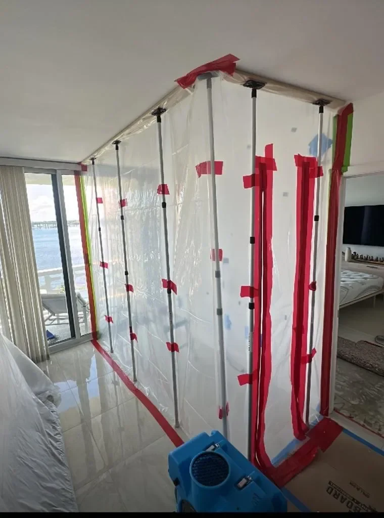 Plastic containment barrier sealed floor to ceiling with poles and tape inside a waterfront condo, with an air mover in front
