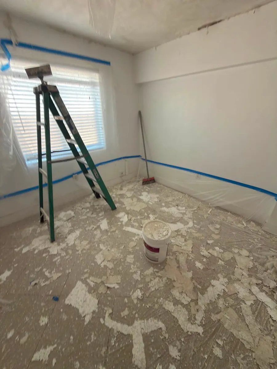 Bedroom during popcorn ceiling removal, with walls and window taped off in plastic and scraped texture on the covered floor