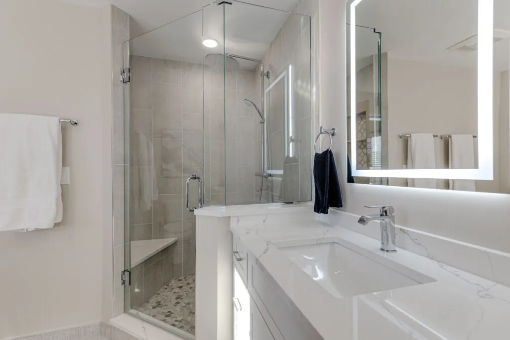 Quartz vanity countertop beside a frameless glass shower with porcelain wall tile, corner bench and pebble floor