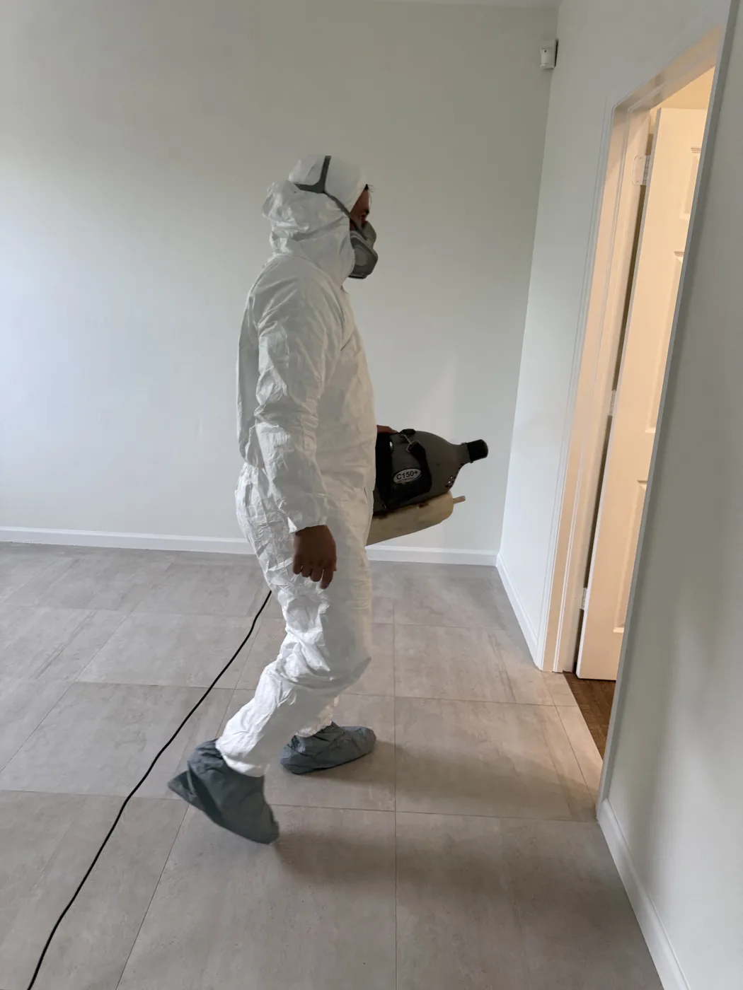 BO Restoration technician in protective suit, respirator, and shoe covers carrying a disinfection fogger through a cleaned room