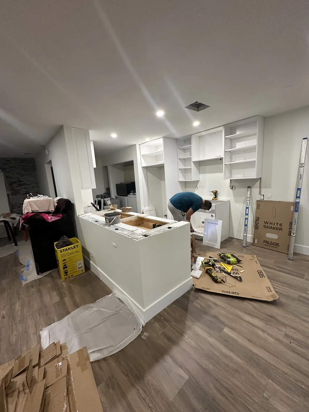 Installer leveling new kitchen base cabinets, with wall cabinet boxes already hung and white shaker doors waiting to go on