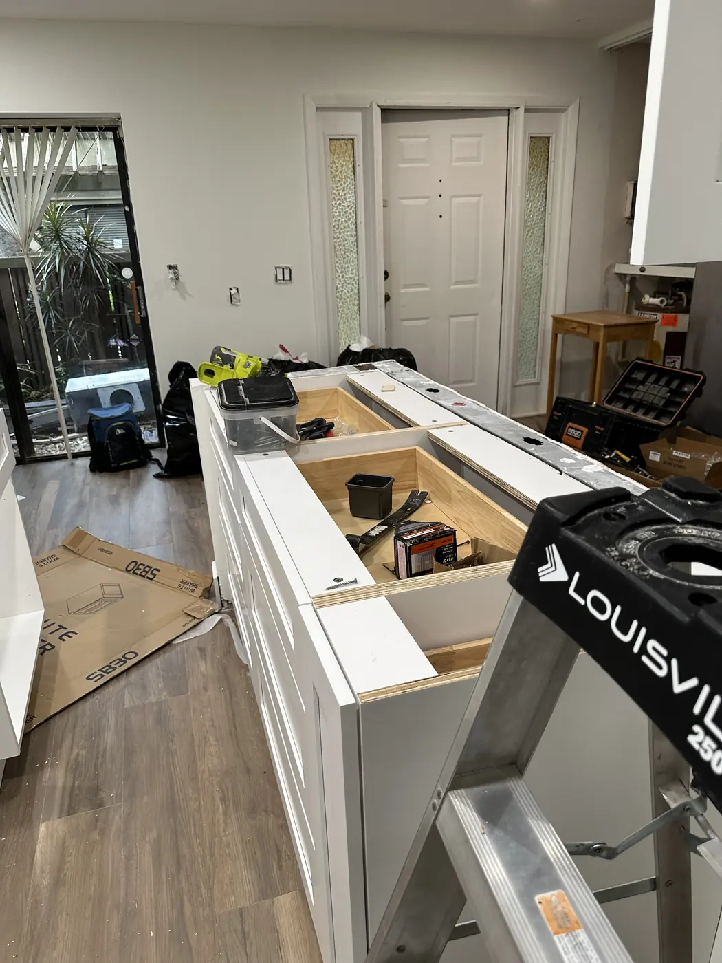 Kitchen island base cabinets being installed, with white shaker drawer fronts and the top open for the countertop