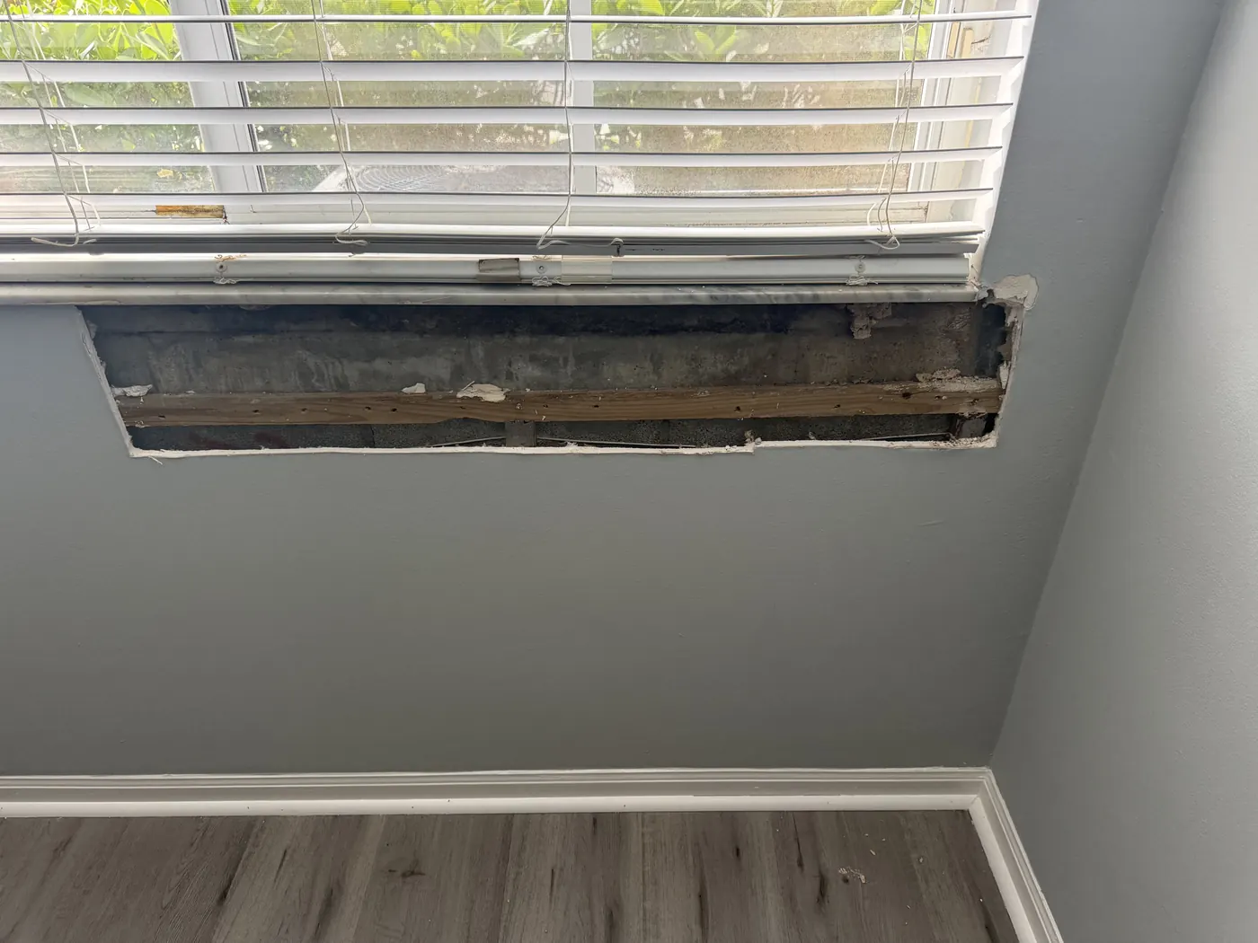 Water-damaged drywall cut out below a window, exposing the block wall and wood furring strip before repair