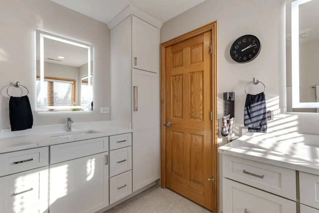 Bathroom with white shaker vanities, a tall shaker linen cabinet and white quartz countertops under backlit mirrors